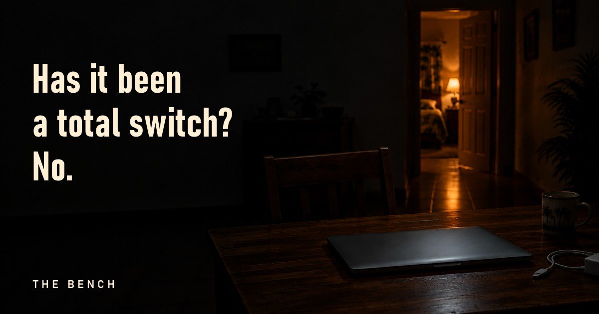 A closed laptop on a dark desk, with warm lamp light from a bedroom door down the hall. Has it been a total switch? No.